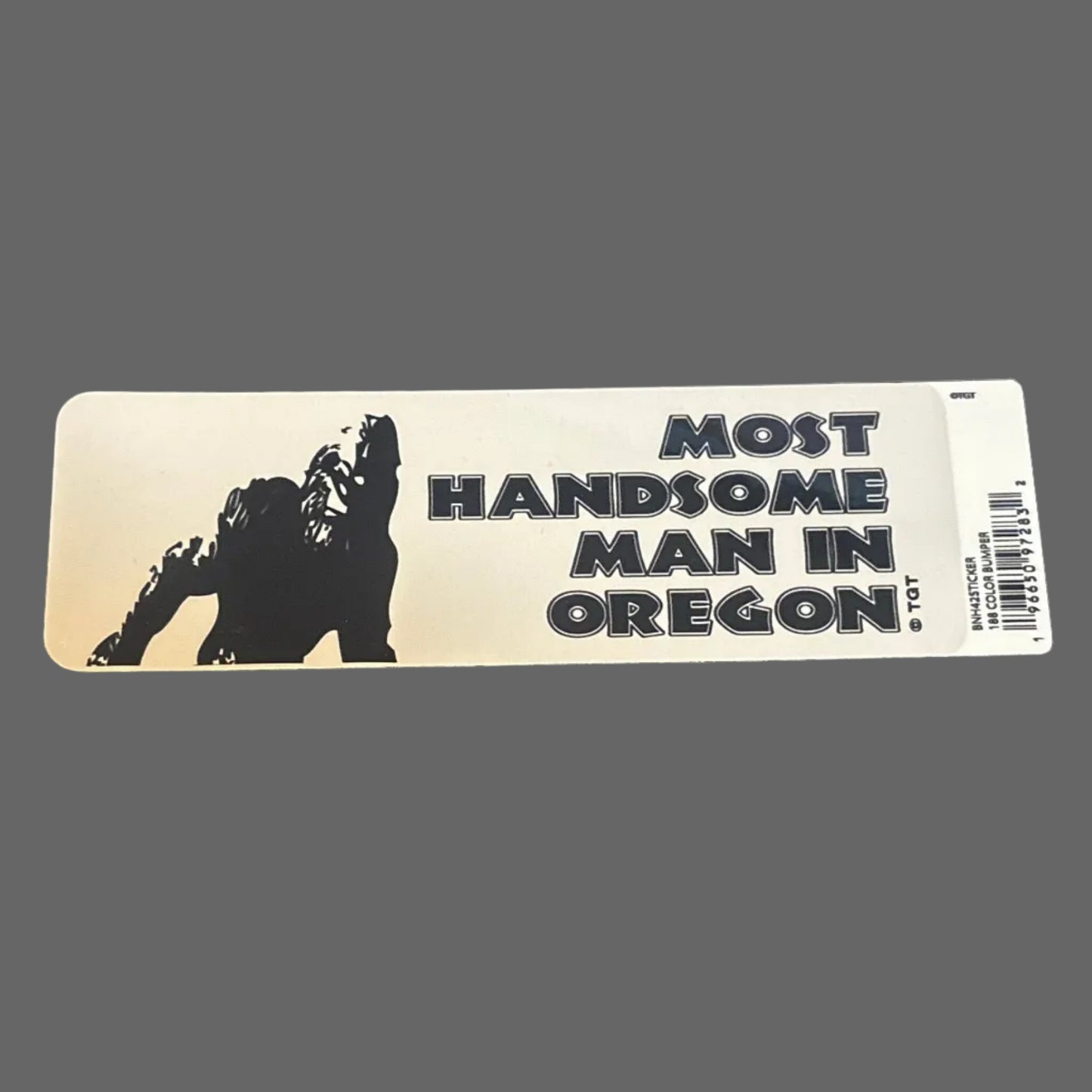 Sticker with 'Most Handsome Man in Oregon' text on a dark surface with a ruler and keyboard in the background.